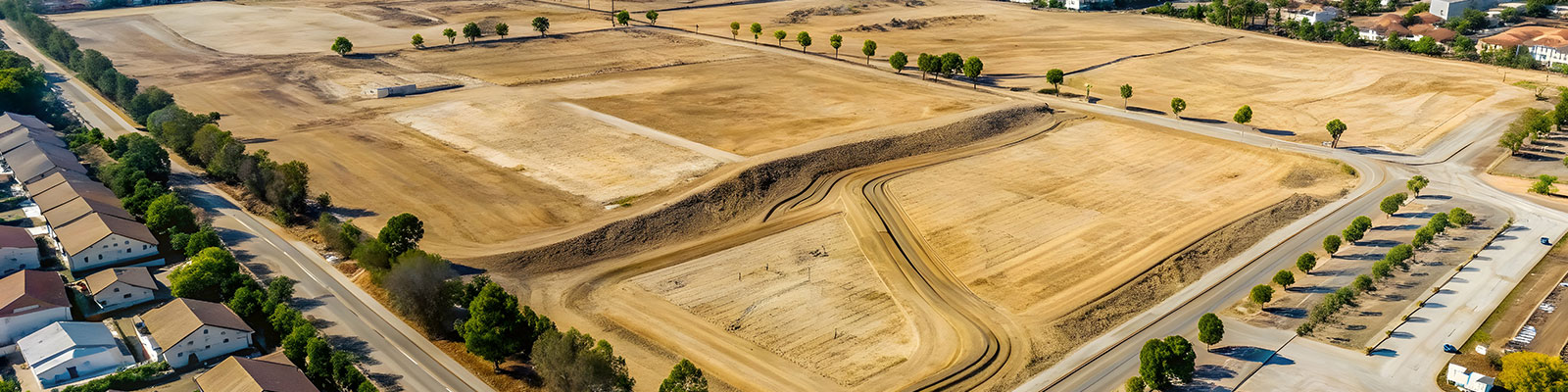 Aerial view of cleared land with dirt roads and leveled plots prepared for future development, bordered by residential homes and lined with trees.