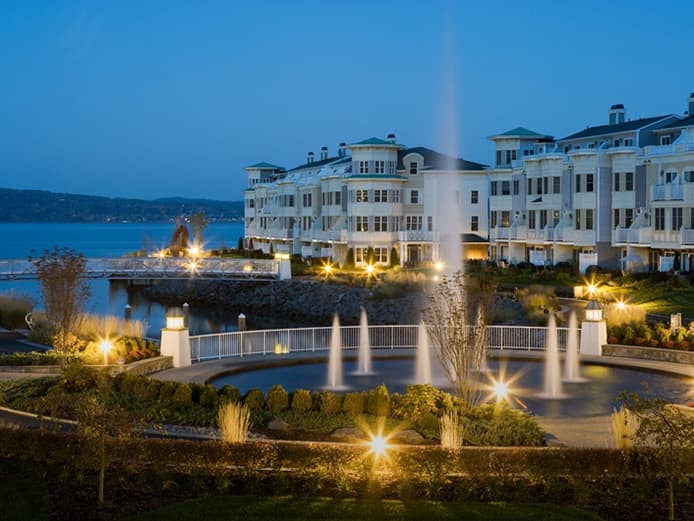 A fountain spraying water with harbor-front homes in the evening.